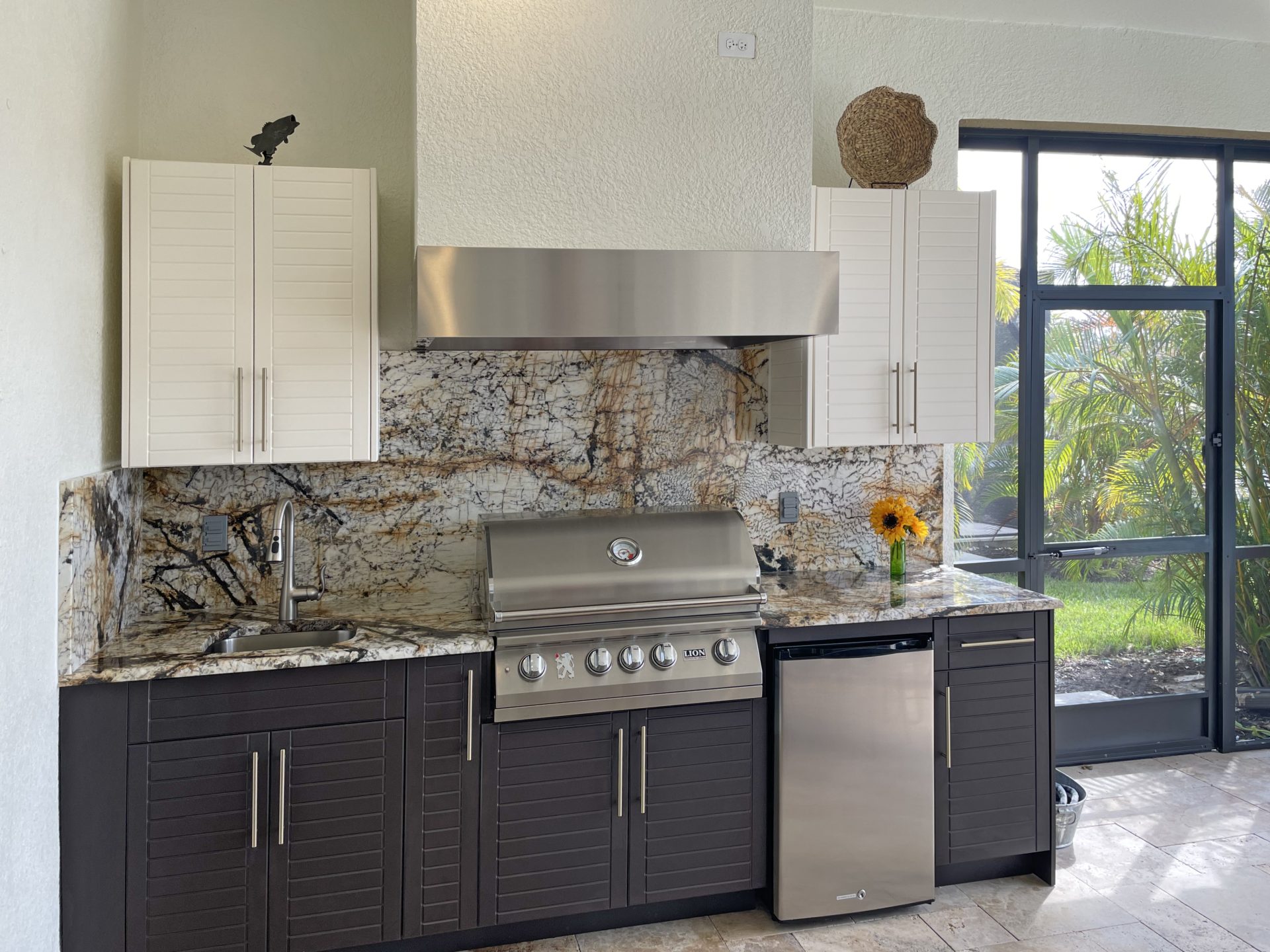 OUTDOOR KITCHEN 17. Custom outdoor kitchen in Nokomis, FL. Kitchen features seafoam and dark brown, cabana cabinets. Appliances include Lion grill, Falmec outdoor hood, stucco hood chase, Blaze stainless front refrigerator and stainless bar pulls.